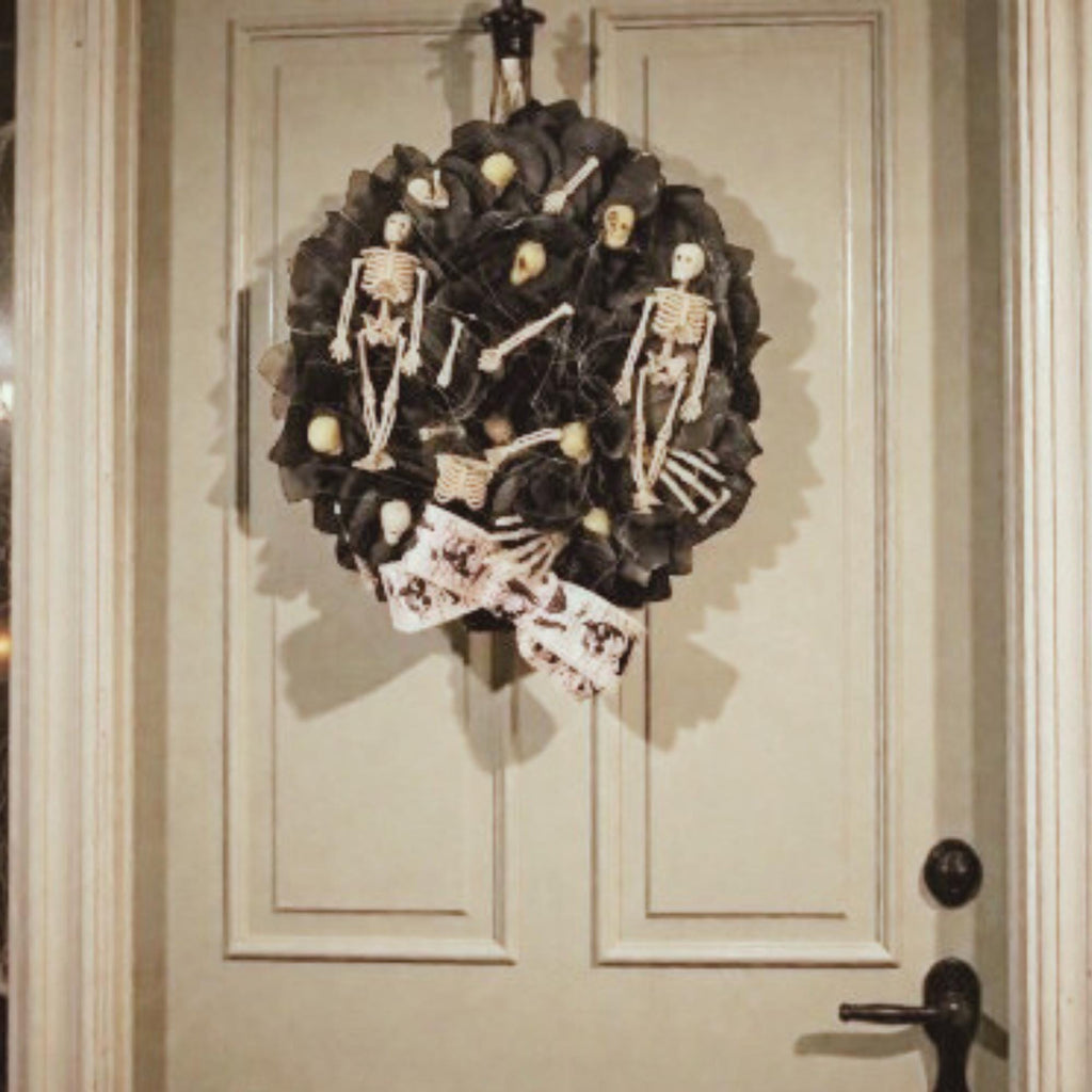 A beautiful round full wreath with a base of black satin & organza flower petals, several tiny skulls, some bone fragments, two skeletons coming out of wreath to hold his bow down and keep him from falling out of the wreath, there are two full skeletons on the sides, and a white satin bow at the bottom with anatomy of bones. it hangs on a modern white door with a black vintage handle