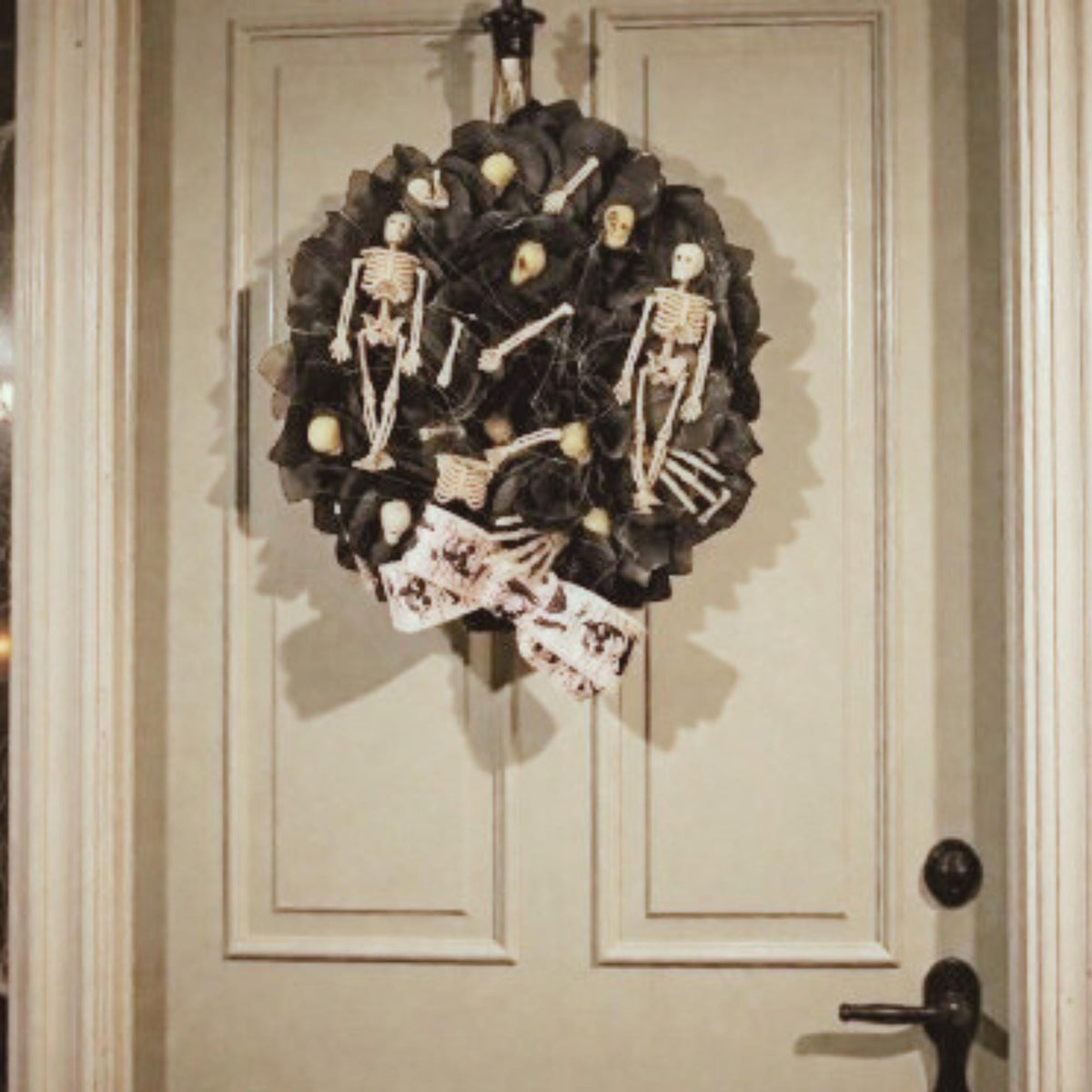 A beautiful round full wreath with a base of black satin & organza flower petals, several tiny skulls, some bone fragments, two skeletons coming out of wreath to hold his bow down and keep him from falling out of the wreath, there are two full skeletons on the sides, and a white satin bow at the bottom with anatomy of bones. it hangs on a modern white door with a black vintage handle